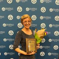 Alum standing in front of the GV Alumni Relations backdrop with an award and flowers.
